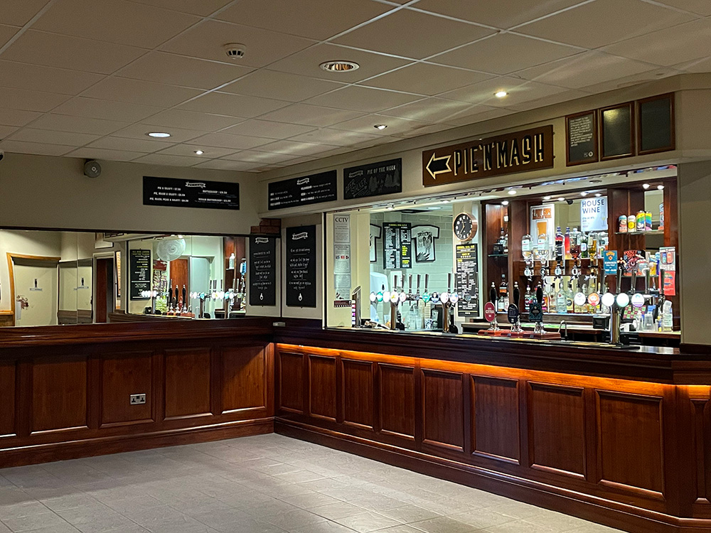 The inside of the main bar room in Brudenell. It is completely empty. On the right is the actual bar with dark brown wood panelling and lots of bottles lining the wall. On the left, there is a mirror.