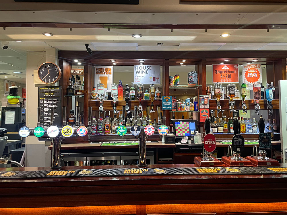 This is a close up of the bar. In the foreground, there are 2 large taps with 5 beer/cider on each. Next to these taps are 3 singular 1-beer taps. The back wall is lined with posters about alcohol deals as well as spirit bottles.