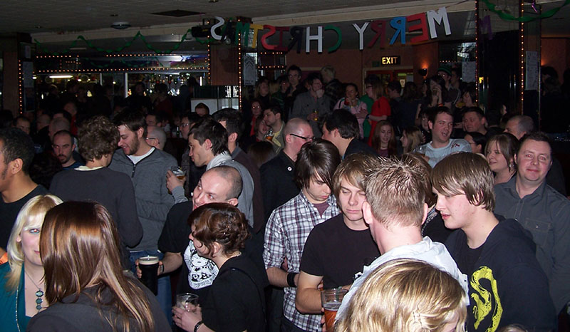 In a dimly lit room, there is a large crowd of people waiting for a gig to commence.