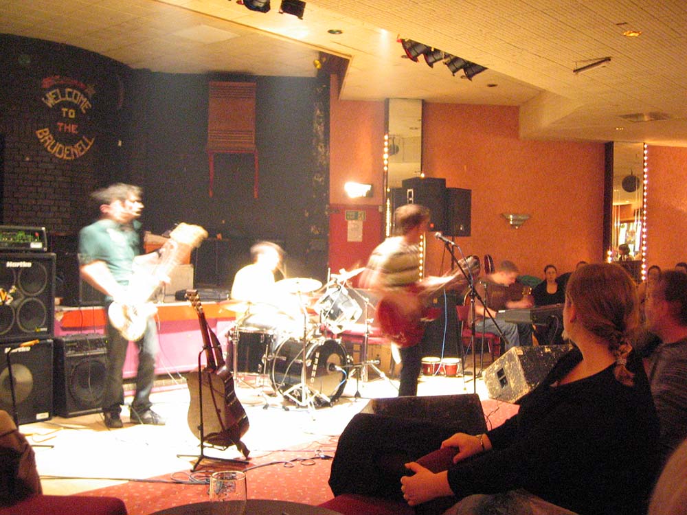A picture of 3 band members. one singing and playing the guitar, another on the drums, and the last playing an electric guitar.