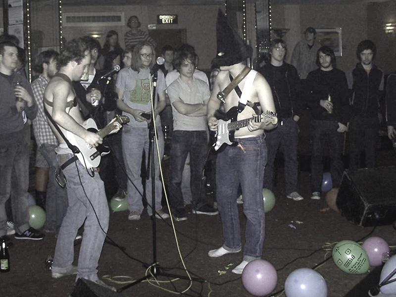 Two band members are playing the electric guitar/bass while a crowd of people stand behind. Theright player is wearing a pointed witch hat covering his whole face.