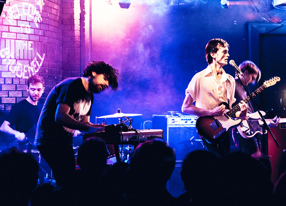 A group of 4 band members are playing a gig amongst a smoky room that has been lit purple and blue.