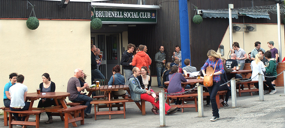 An shot of the outside of Brudenell Social Club. 4 benches line the building and there are lots of people sat on them drinking pints of beer.
