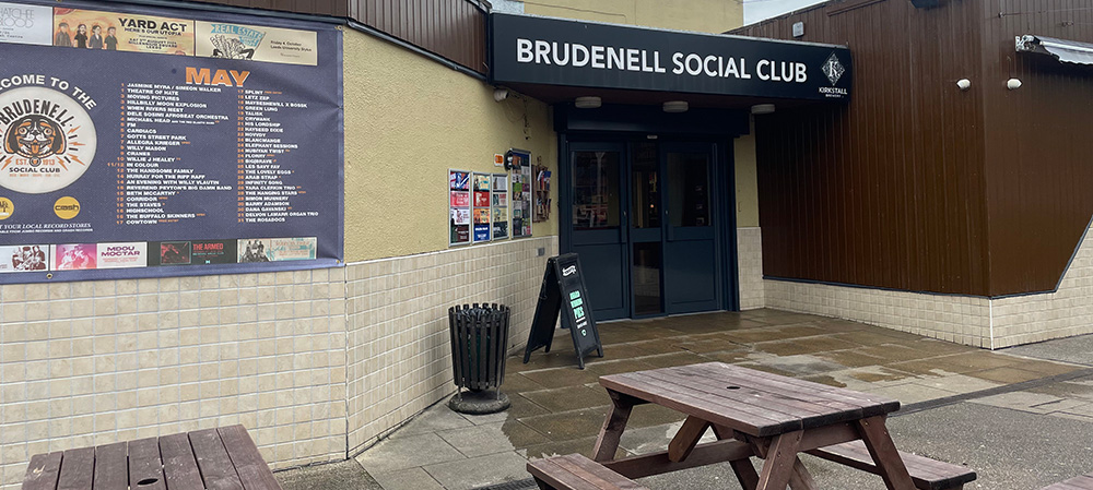 At the same angle as the above image, this image shows the outside of Brudenell but empty. On the right is the building's sign and entrance door and on the left shows a large poster covering the wall stating the list of gigs for the month of may