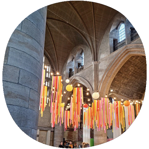 The interior of an old church with yellow, white, and orange lanterns. There are colourful streamers coming from the top.