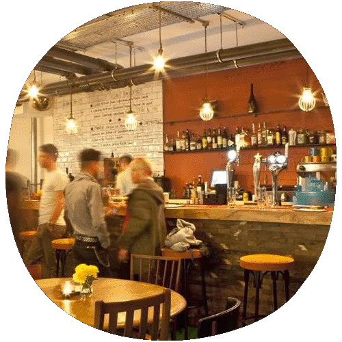 Brightly lit bar with blurred figures passing. The wall behind the bar is painted red while the rest of the tiles are white. The back wall is lined by bottles. 