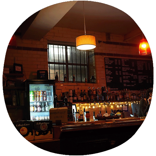 Dimly lit bar at Wharf Chambers. There are rows of bottles with fairy lights underneath. On the left there is a fridge with a transparent door.