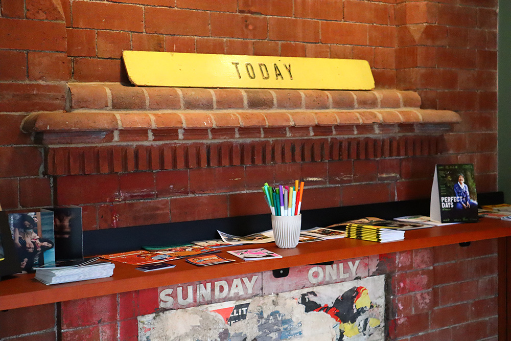 A red bricked wall above where a fireplace would go. At the top of the brick wall shelf, there is a yellow sign that says today. Beneath there is a white pencil pot and lots of flyers.