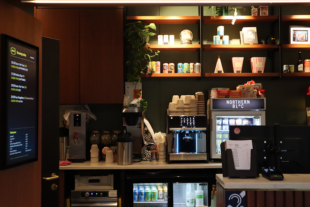 It shows Hyde Park Picture Houses's snack bar and ticket shop. In the foreground there is a till and in the background, cans of drinks, empty popcorn boxes, and a coffee machine line a dark green wall.