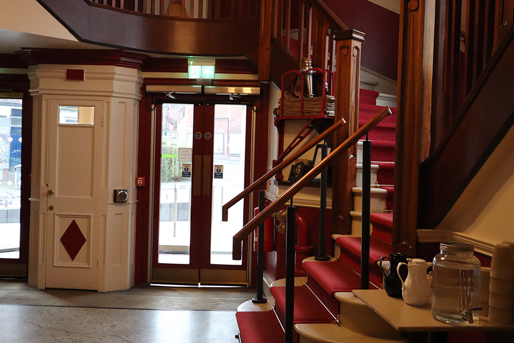It depicts the double door entrance to the cinema lined with red paint. On the right side of the image is the white grand staircase that has red carpet lining.