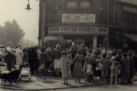 The image shows a crowd of people around an elephant. It is taken in front of hyde park picture house's entrance. At the back of the crowd, there seem to be lots of mothers holding babies with prams.