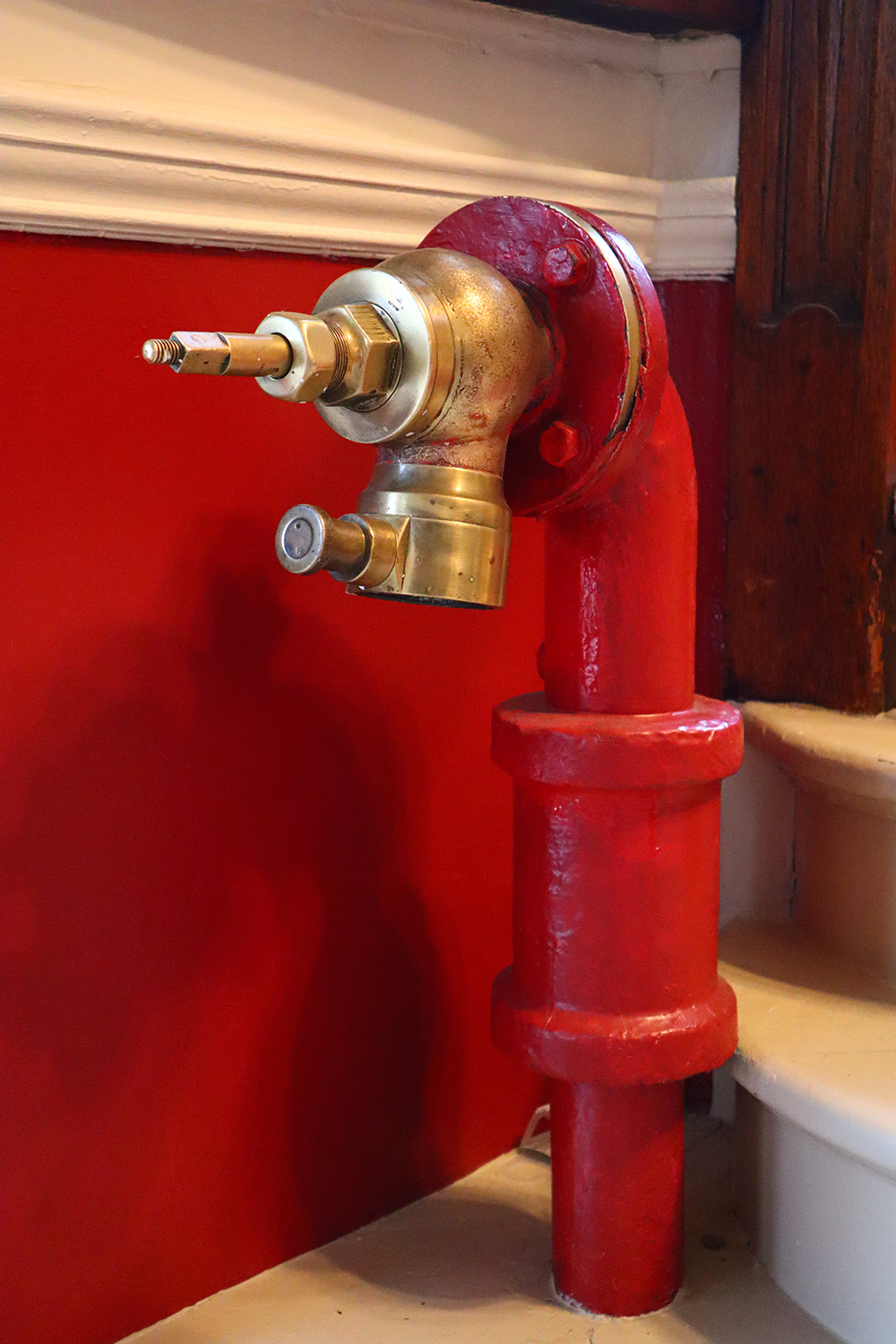 An image of the full old brass fire hydrant. The base of the hydrant is HPPH red while the spout is a golden brass colour. It stands against a red wall with white trim.