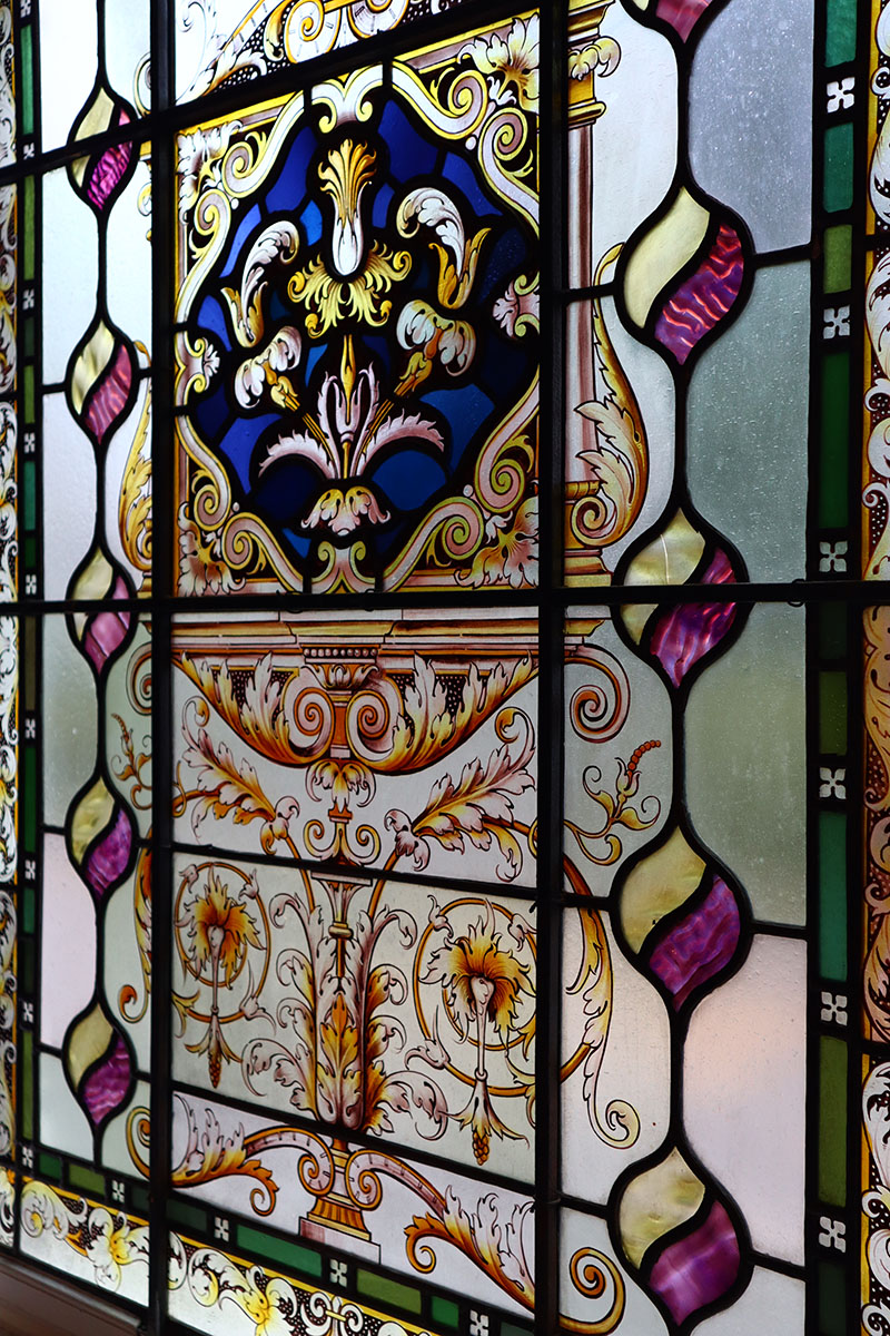 A close up of the stained glass window taken from a 45 right angle. The stained glass is lined with an alternating dark/light green border. Moving further inwards, it shows repeated lemon shapes that ascend down the window, halved with a yellow and purple colour. Further inwards shows intricate golden lines to make up the middle section