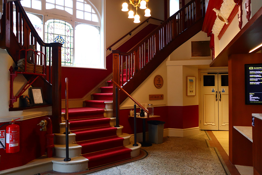 A wide angle of the grand staircase with dark brown wood railings. Behind the white staircase lined with plush red fabric is the stained glass.