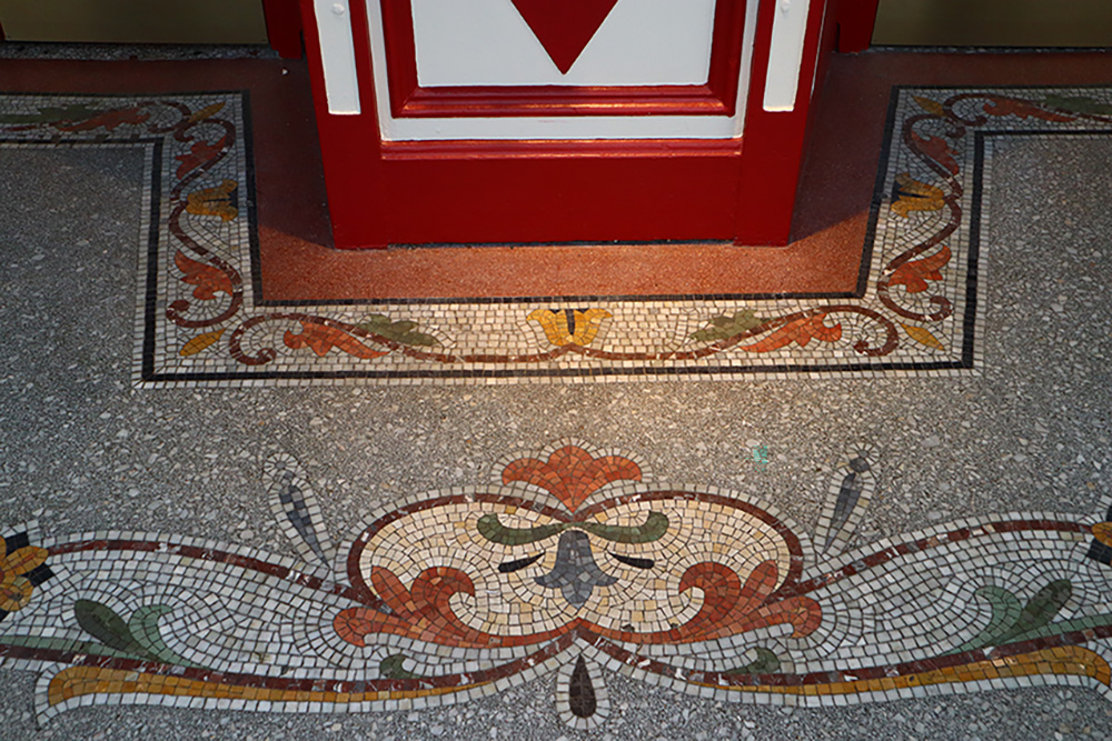 A downward shot to see the tiled floor just outside the ticket booth area. The base colour are different variations of grey tiles while a large detail runs horizontally across the floor. The detail involves terracotta, wallflower yellow, denim blue, and forest green tiles assembled in curving shapes.