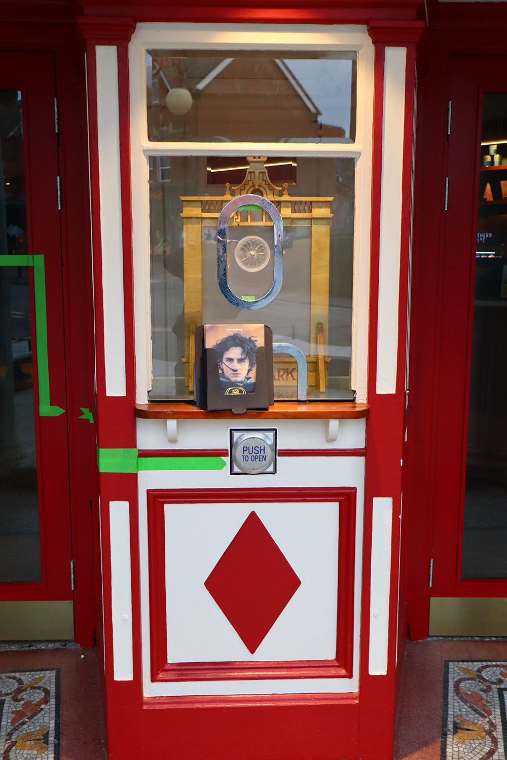 A portrait image of the traditional cinema ticket booth. The white box is decorated with dark red lines and diamond shapes. Sitting in front of the ticket booth is a copy of the monthly film schedule. The cover depicts actor Timothee Chalamet in Dune Part Two.