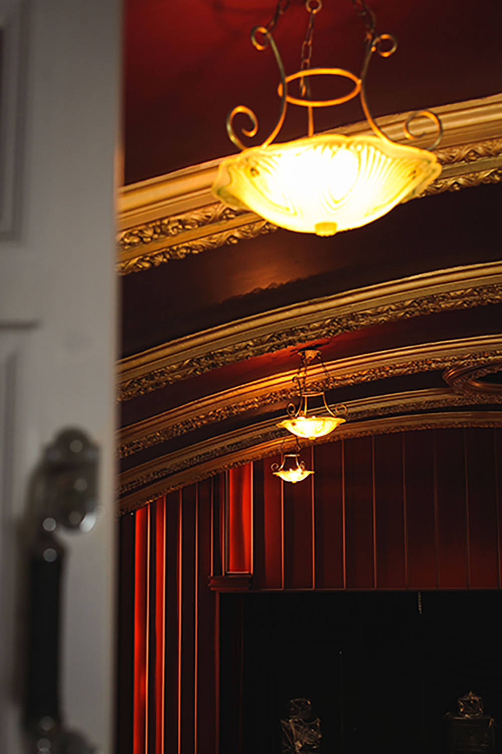 An image focusing on the ceiling gas lamp on the upper floor of the main cinema room. It shows a blurred open door in the foreground and a row of 3 gas lamps going further away from the camera