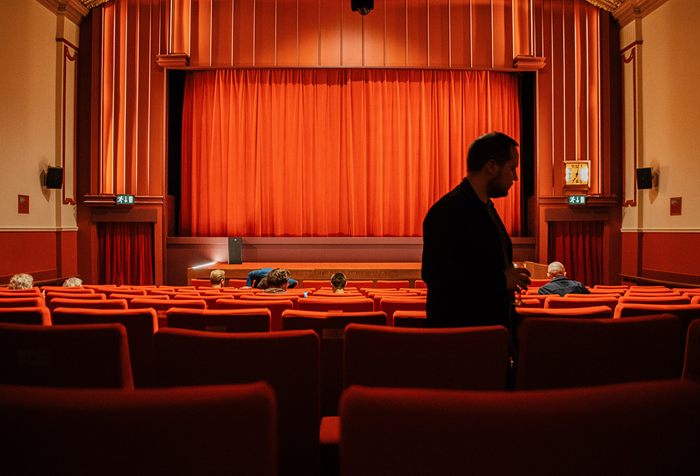 Showing the same subject as the image above but in colour, this image shows the red curtains once again covering the screen. Taken from the bottom floor, there are lots of seats in the foreground with a man walking to the right of the image.