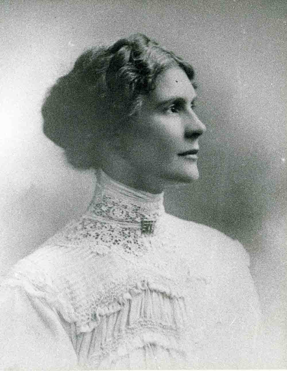 A portrait image of Leonora Cohen. She is facing towards the right side of the photo with a neutral expression. Her hair is braided around her head and she wears a lace decorated top or dress.